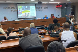 Foto de um auditório da OAB-MG com mulheres e homens acompanhando uma palestra com uma apresentação projetada em um telão.