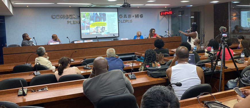 Foto de um auditório da OAB-MG com mulheres e homens acompanhando uma palestra com uma apresentação projetada em um telão.