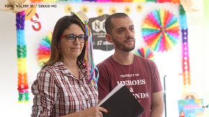 Janaina Torres, diretora de aposentados e pensionistas, vestindo uma camisa xadres e um pequeno chapeu com fitas coloridas no cabelo. Ao seu lado, está Alexandre Pire, coordenador Geral do SINJUS, vestindo uma camisa vinho. Ao fundo, é possível ver enfeites coloridos alusivos à festa junina, 