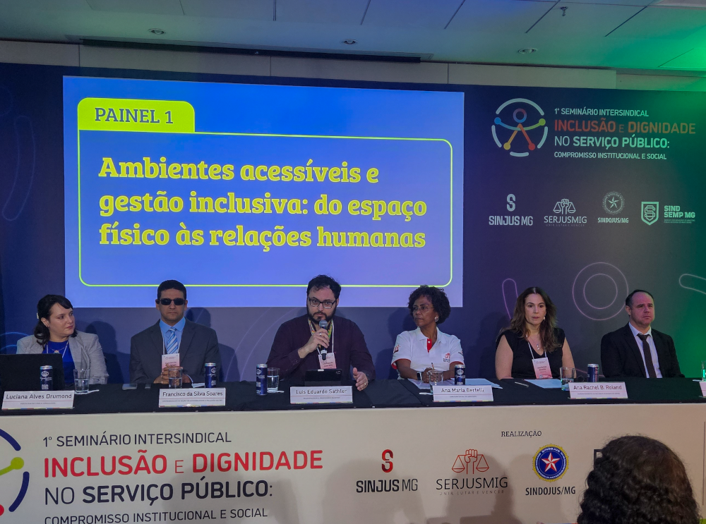 Em um auditório, seis pessoas estão sentadas lado a lado em uma mesa retangular, compondo o Painel 1 do 1º Seminário Intersindical de Inclusão e Dignidade no Serviço Público. Ao fundo, em um telão azul com letras amarelas, está escrito o título do painel: “Ambientes acessíveis e gestão inclusiva: do espaço físico às relações humanas”. Da esquerda para a direita: Luciana Alves Drummond, mulher de pele clara, cabelos castanhos presos, veste blazer cinza sobre blusa azul e observa com atenção. Francisco da Silva Soares, homem de pele clara, usa óculos escuros, terno cinza e gravata azul clara, com expressão séria e postura ereta. Luiz Eduardo Sathler, homem de pele clara, barba cheia e óculos, veste camisa azul marinho e fala ao microfone com gesto de envolvimento. Ana Maria Bertelli, mulher negra, cabelos crespos curtos, veste blusa branca, com postura firme e expressão acolhedora. Ana Rachel B. Roland, mulher de pele clara, cabelos castanhos soltos, veste colete preto sobre blusa branca, olhando com atenção para a plateia. Willian Nascentes, homem de pele clara, cabelos curtos, veste terno escuro e gravata preta, sentado de forma formal e atenta.