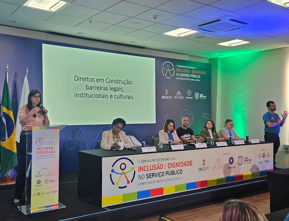Foto do 1º Seminário Intersindical Inclusão e Dignidade no Serviço Público. À esquerda, Kátia Roncada, mulher de cabelos castanhos e óculos, fala ao microfone atrás de um púlpito. Sentados à mesa estão Cleonice Amorim, mulher negra de cabelos curtos e óculos; Fanny Melo, mulher de cabelos castanhos lisos; Wagner Ferreira, homem de barba com camiseta preta; Paloma Resende, mulher de cabelos longos castanhos; e Flávia Felício, mulher de cabelos presos e óculos. À direita, um intérprete de Libras, homem de barba e camiseta azul, faz a tradução.