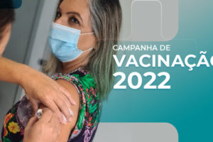 Foto de uma mulher de pele clara, loira, usando máscara com um vestido estampado, em pé, recebendo sua dose da vacina contra a gripe. Na parte inferior em detalhes azuis temos o conteúdo textual " Campanha de vacinação 2022"