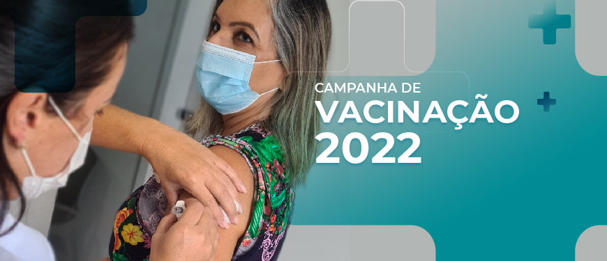 Foto de uma mulher de pele clara, loira, usando máscara com um vestido estampado, em pé, recebendo sua dose da vacina contra a gripe. Na parte inferior em detalhes azuis temos o conteúdo textual " Campanha de vacinação 2022"