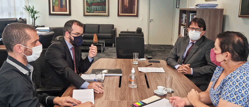 A imagem contém quatro pessoas reunidas em uma mesa de madeira, sendo que, sentados de um lado os dirigentes do SINJUS Alexandre Pires e Felipe Rodrigues, e do outro 1º vice-presidente do TJMG, desembargador Alberto Vilas Boas, e com a juíza auxiliar da 1ª Vice-Presidência, Mônica Silveira Vieira. No fundo da imagem existe uma parede branca com cinco quadros emoldurados em madeira um conjunto de sofás na cor preta. 