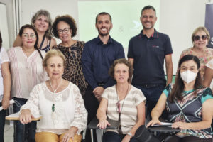 A imagem contém um grupo de treze pessoas sorrindo, sendo onze mulheres e dois homens. Ao fundo há uma parede branca com um telão e um banner do Sinjus MG. Posicionadas à frente do grupo estão quatro mulheres sentadas em suas respectivas cadeiras.