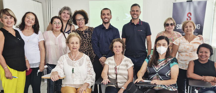 A imagem contém um grupo de treze pessoas sorrindo, sendo onze mulheres e dois homens. Ao fundo há uma parede branca com um telão e um banner do Sinjus MG. Posicionadas à frente do grupo estão quatro mulheres sentadas em suas respectivas cadeiras.